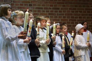 Erstkommunionkinder in Anzuegen und weissen Kleidern halten brennende Kommunionkerzen in ihren Haenden