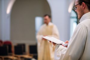 Ein Kantor in liturgischer Kleidung singt, im Hintergrund ein Priester