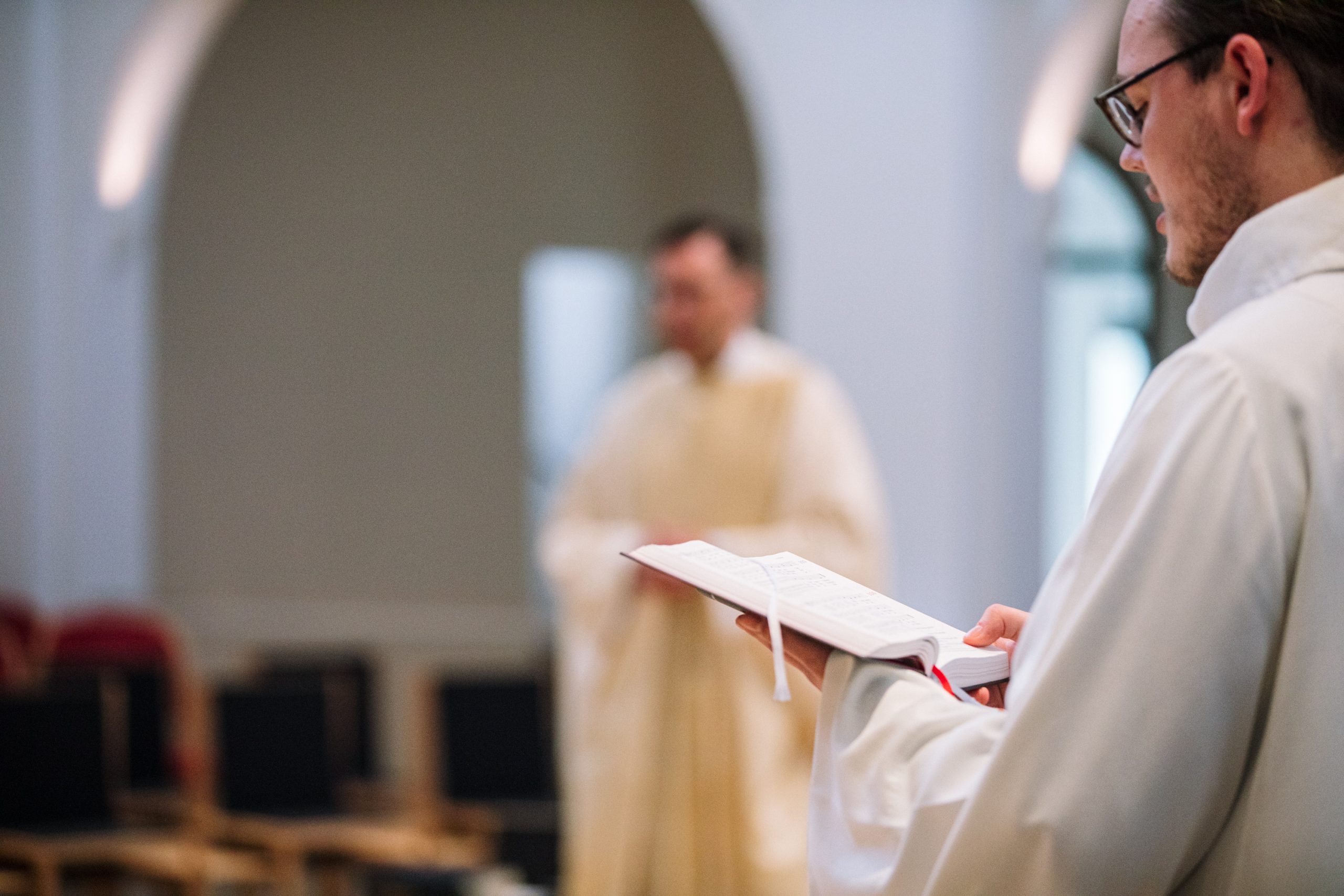 Ein Kantor in liturgischer Kleidung singt, im Hintergrund ein Priester