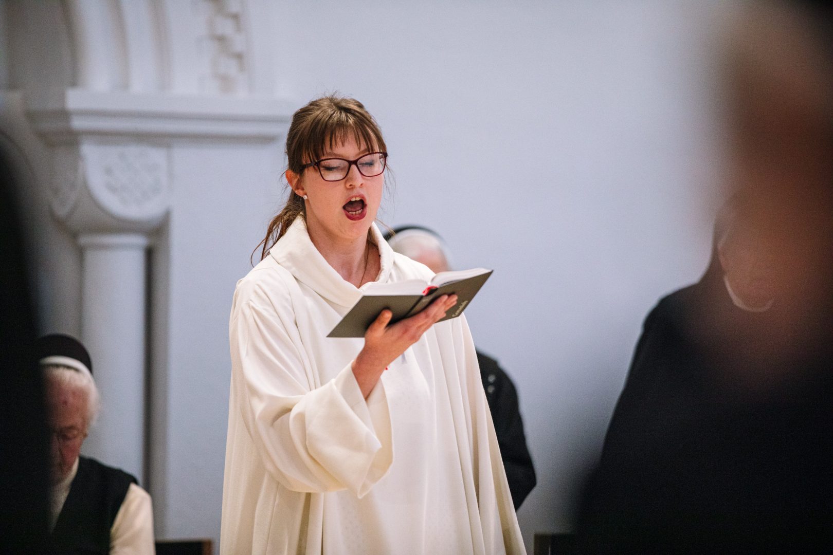 Eine Kantorin in liturgischer Kleidung singt, im Hntergrund Ordensschwestern