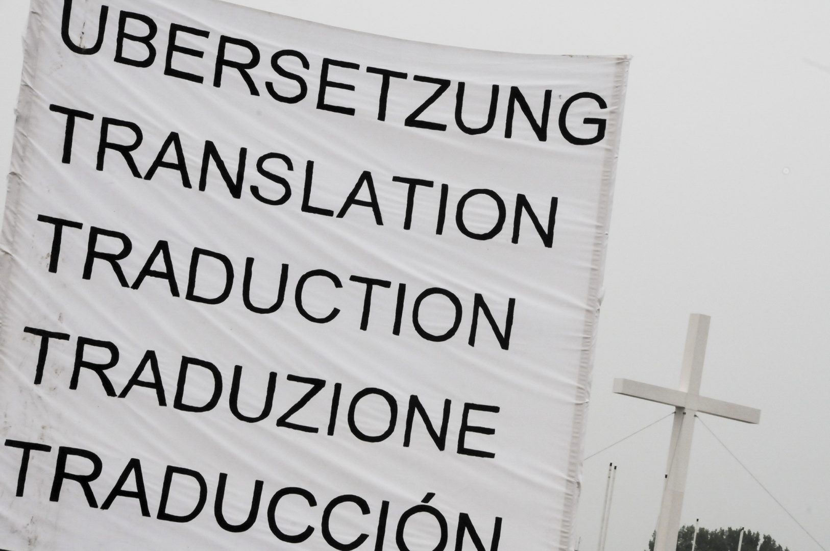 Ein Transparent mit dem Titel Übersetzung auf verschiedenen Sprachen, im Hintergrund ein weißes Kreuz