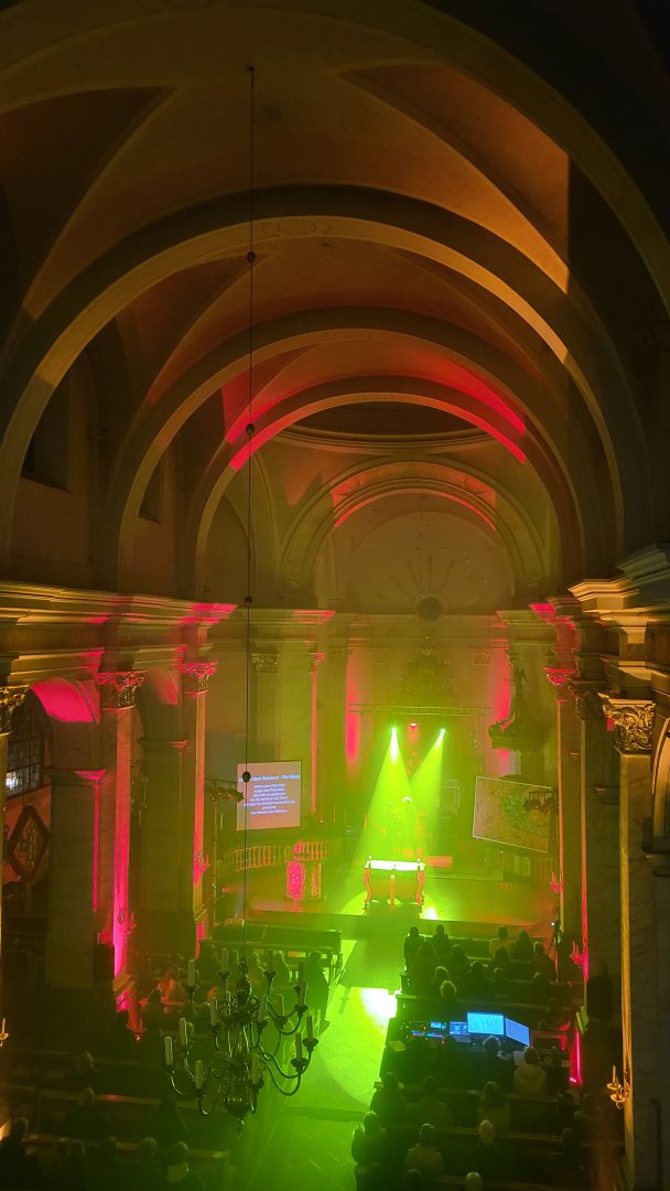 Kirchenschiff mit buntem Licht, gelb und rosa Strahler, junge Menschen sitzen in den Kirchenbaenken